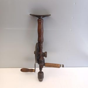 Op de afbeelding: Een vintage handboor met een verticaal handvat van donkerbruin hout met een metalen bovenplaat, een centraal tandwielmechanisme, een zijwaartse zwengel met een houten knop, een horizontaal houten handvat en een metalen boorkop aan de onderzijde.