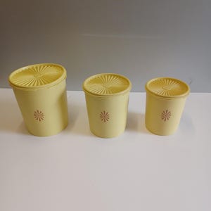 Könnte beinhalten: Drei gelbe Vintage-Lebensmittelbehälter mit passenden Deckeln. Jeder Behälter hat ein Blumenmuster in der Mitte. Die Deckel haben ein radiales Design. Die Behälter sind in verschiedenen Größen.
