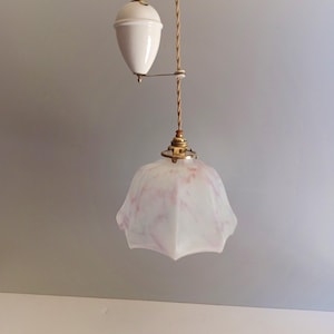 Może przedstawiać: Wisząca lampa z kloszem z matowego szkła w kształcie dzwonu z delikatnymi różowymi marmurkowymi wzorami. Lampa jest zawieszona na skręconym sznurku z tkaniny połączonym z systemem przeciwwagi z białą ceramiczną rolką i mosiężnymi okuciami.