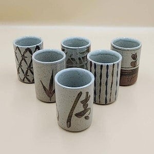 May include: Six ceramic cups with various designs. The cups are cylindrical with a speckled, off-white base color and a light blue interior. Designs include brown geometric patterns, vertical stripes, and Japanese calligraphy.