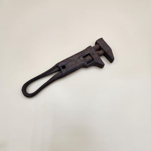 May include: A rusty, vintage adjustable wrench. The wrench is dark brown with a loop handle and a jaw that can be adjusted. The word "ROSE MFG" is stamped on the wrench.