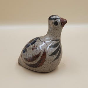 May include: A ceramic bird figurine with a speckled gray body, featuring painted details in blue, brown, and black. The bird has a dark gray head with a brown beak and a large, round eye. Decorative art piece.