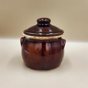 May include: A small, brown ceramic pot with a lid. The pot has a glossy finish and two small handles. The lid is slightly darker than the body of the pot and has a small knob on top. The rim of the pot is a lighter color.