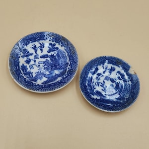May include: Two antique blue and white porcelain saucers with a Willow pattern design. The saucers feature detailed scenes of a bridge, pagoda, and figures. One saucer has a small chip on the rim.