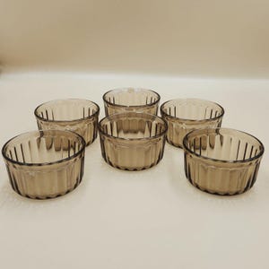 May include: Six clear, brown-tinted glass ramekins. Each ramekin has a round shape with vertical ridges on the sides. The ramekins are arranged in a group, suitable for baking and serving individual portions of food.