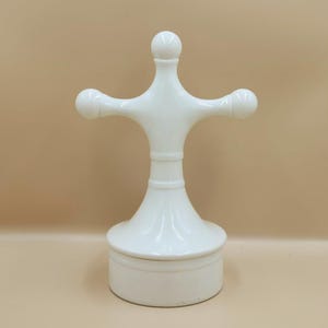 May include: A white, decorative bottle stopper with a unique design. The stopper features a cylindrical base, a flared midsection, and a top with a cross-like shape, with rounded ends. The overall design is smooth and glossy.