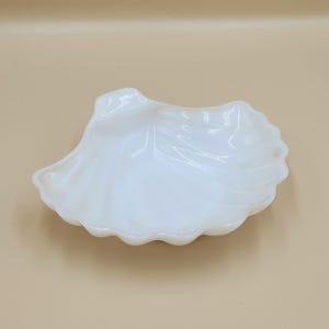 May include: A white, seashell-shaped dish with a scalloped edge. The dish has a smooth, glossy surface and is likely made of ceramic or glass. The dish is empty and sits on a neutral background. This decorative dish is perfect for holding small items.