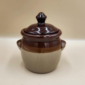 May include: A small, ceramic jar with a lid. The jar is a light beige color on the bottom, with a brown band in the middle and a dark brown lid. It has two small handles on the sides.
