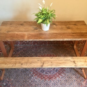 May include: A rustic wooden dining table and bench set with copper-colored metal legs. The table and bench have a natural wood finish and are set on a patterned rug.