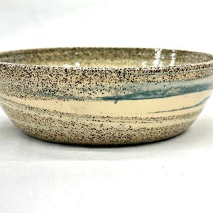 May include: A round ceramic bowl featuring a speckled beige and brown exterior with horizontal streaks of muted blue glaze. The bowl has a wide, open top and a smooth, curved silhouette, showcasing a textured finish across the entire surface.