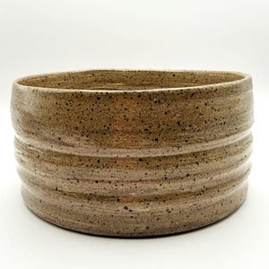 May include: A cylindrical ceramic bowl with a speckled tan and brown finish. The exterior surface features three distinct horizontal ridges circling the body, creating a textured appearance with a smooth, glossy glaze over the speckled clay base.