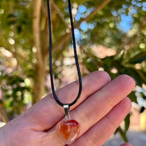 May include: A heart-shaped carnelian pendant on a black cord necklace. The carnelian is a translucent orange-red color with white streaks.
