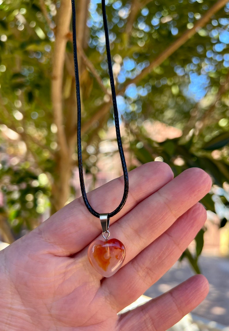 May include: A heart-shaped carnelian pendant on a black cord necklace. The carnelian is a translucent orange-red color with white streaks.