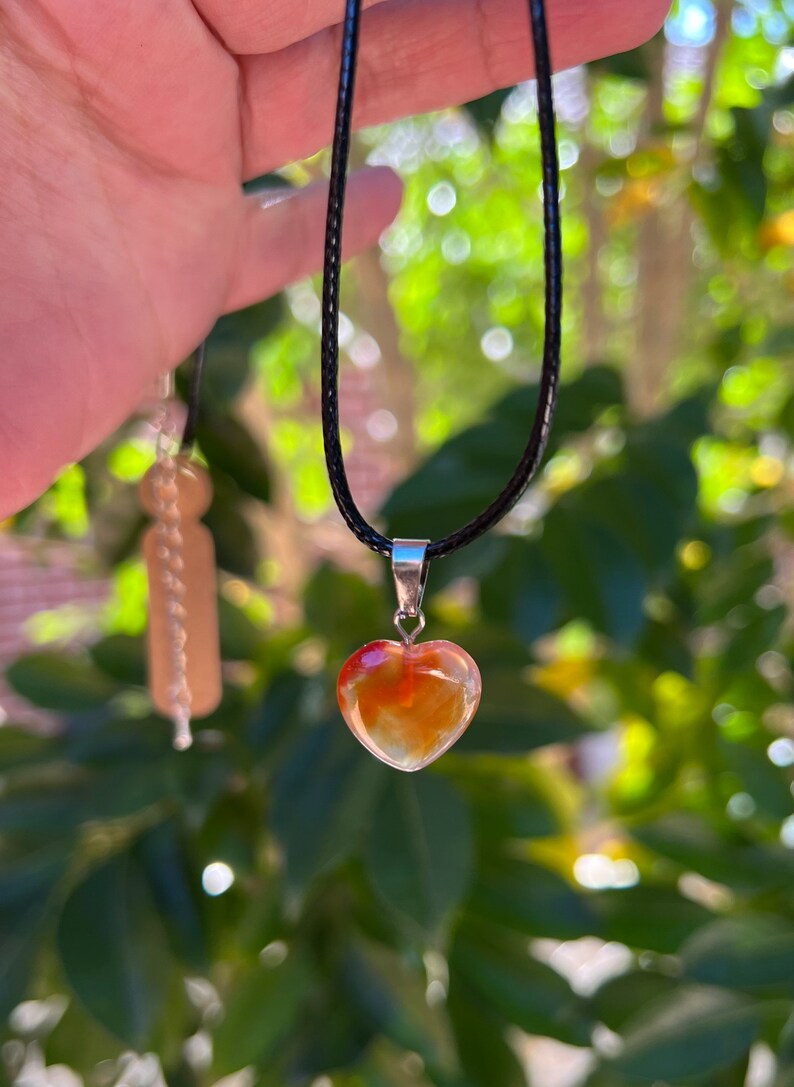 May include: A heart-shaped carnelian pendant on a black cord necklace. The pendant is a light orange color with red and yellow streaks.
