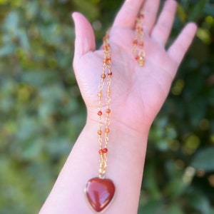 May include: A gold chain necklace with a heart-shaped carnelian pendant. The carnelian is a deep red color and the chain is made of small gold beads.