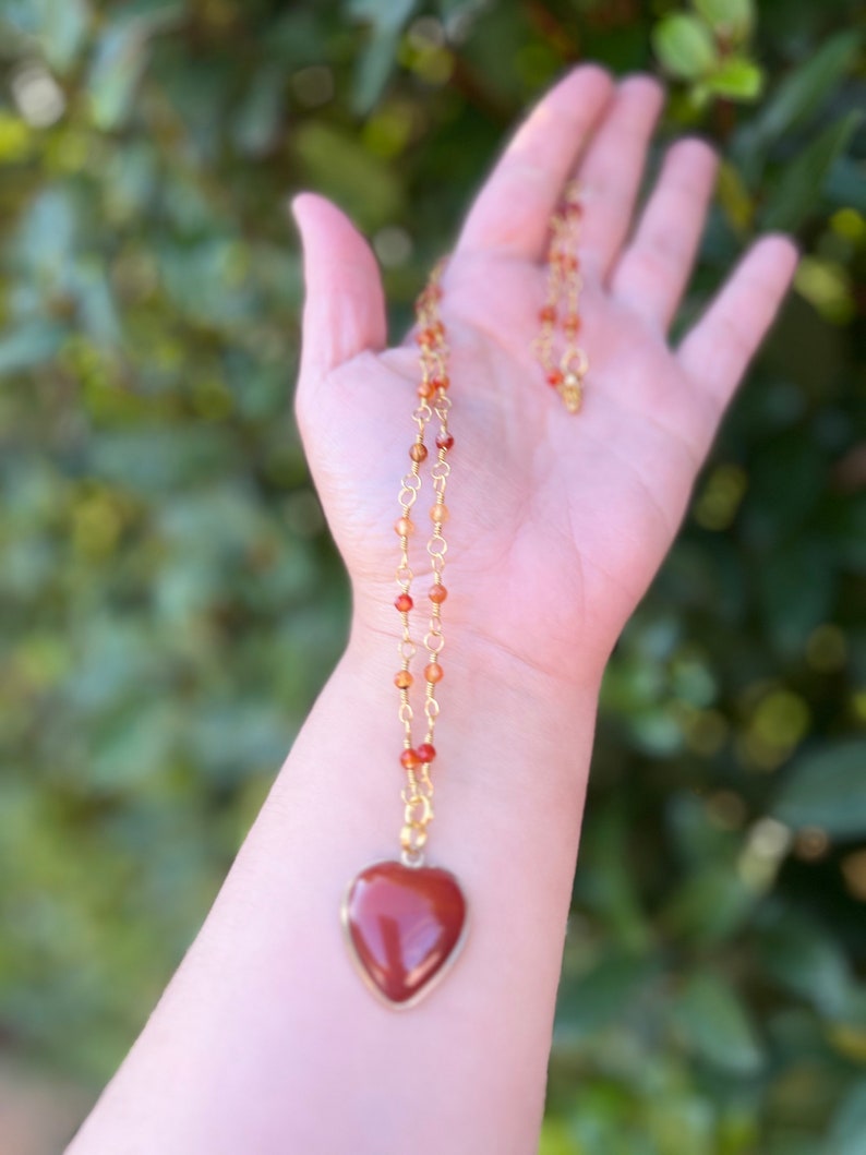 May include: A gold chain necklace with a heart-shaped carnelian pendant. The carnelian is a deep red color and the chain is made of small gold beads.