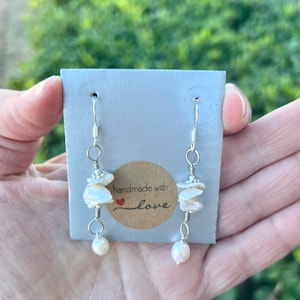 May include: A pair of silver dangle earrings with white pearl accents and a stack of white shell beads. The earrings are displayed on a white card with a brown tag that reads "handmade with love".
