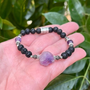 May include: A bracelet with black lava rock beads, silver-toned metal spacers, and small purple amethyst beads. A large, raw amethyst stone is the central feature. The bracelet is set against a backdrop of green foliage.