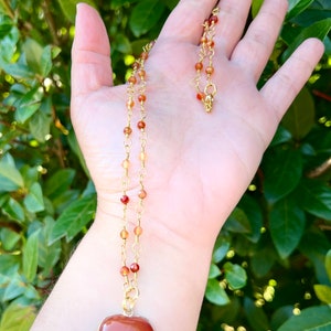 May include: A gold chain necklace with a heart-shaped carnelian pendant. The necklace is made with small carnelian beads and gold chain links.