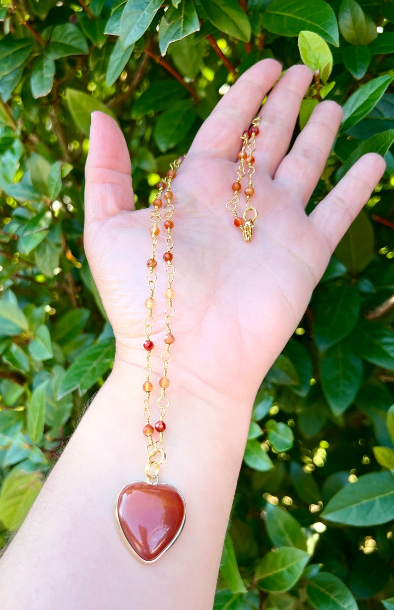 May include: A gold chain necklace with a heart-shaped carnelian pendant. The necklace is made with small carnelian beads and gold chain links.