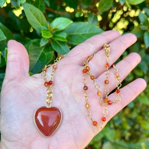 May include: A gold chain necklace with a heart-shaped carnelian pendant and carnelian beads. The necklace is on a hand against a green leafy background.