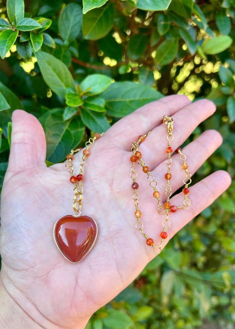 May include: A gold chain necklace with a heart-shaped carnelian pendant and carnelian beads. The necklace is on a hand against a green leafy background.