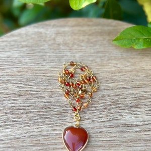 May include: A gold chain necklace with a heart-shaped carnelian pendant. The chain is made of small gold beads and carnelian stones.