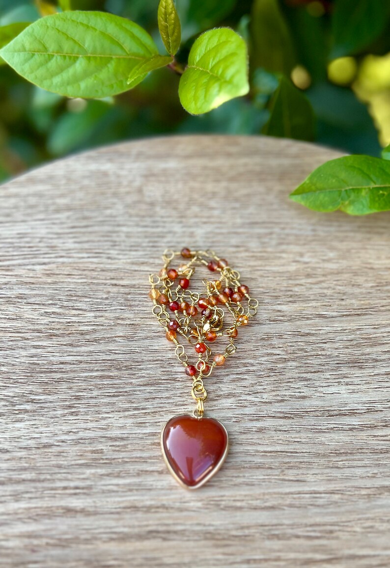May include: A gold chain necklace with a heart-shaped carnelian pendant. The chain is made of small gold beads and carnelian stones.
