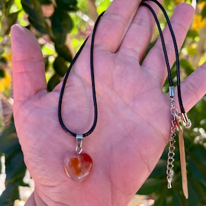 May include: A heart-shaped carnelian pendant on a black cord necklace. The pendant has a translucent orange and red color with white veins.