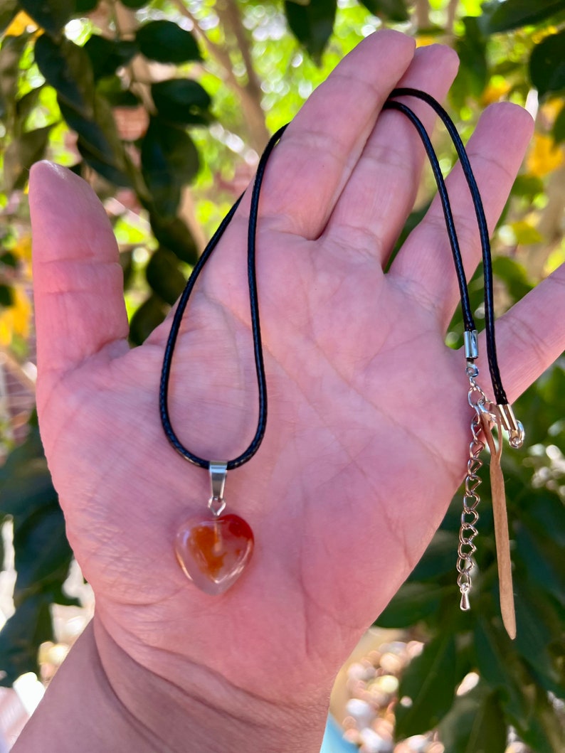 May include: A heart-shaped carnelian pendant on a black cord necklace. The pendant has a translucent orange and red color with white veins.