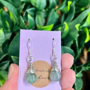 May include: Handmade earrings featuring pale green, teardrop-shaped stones encased in silver wire. The earrings are suspended from silver hooks. A purple card with the words "edmade with love" is in the background.