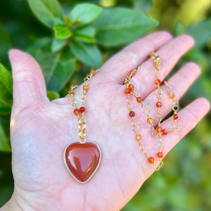 May include: A gold chain necklace with a heart-shaped carnelian pendant. The chain is made of small gold beads and wire.