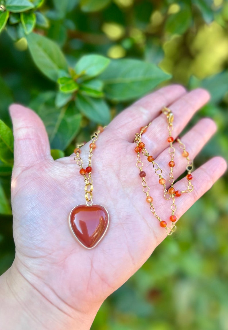 May include: A gold chain necklace with a heart-shaped carnelian pendant. The chain is made of small gold beads and wire.