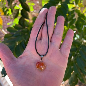 May include: A heart-shaped carnelian pendant necklace on a black cord. The carnelian is a deep orange color with white and brown swirls.