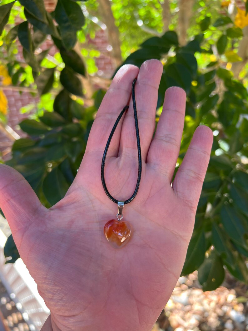 May include: A heart-shaped carnelian pendant necklace on a black cord. The carnelian is a deep orange color with white and brown swirls.