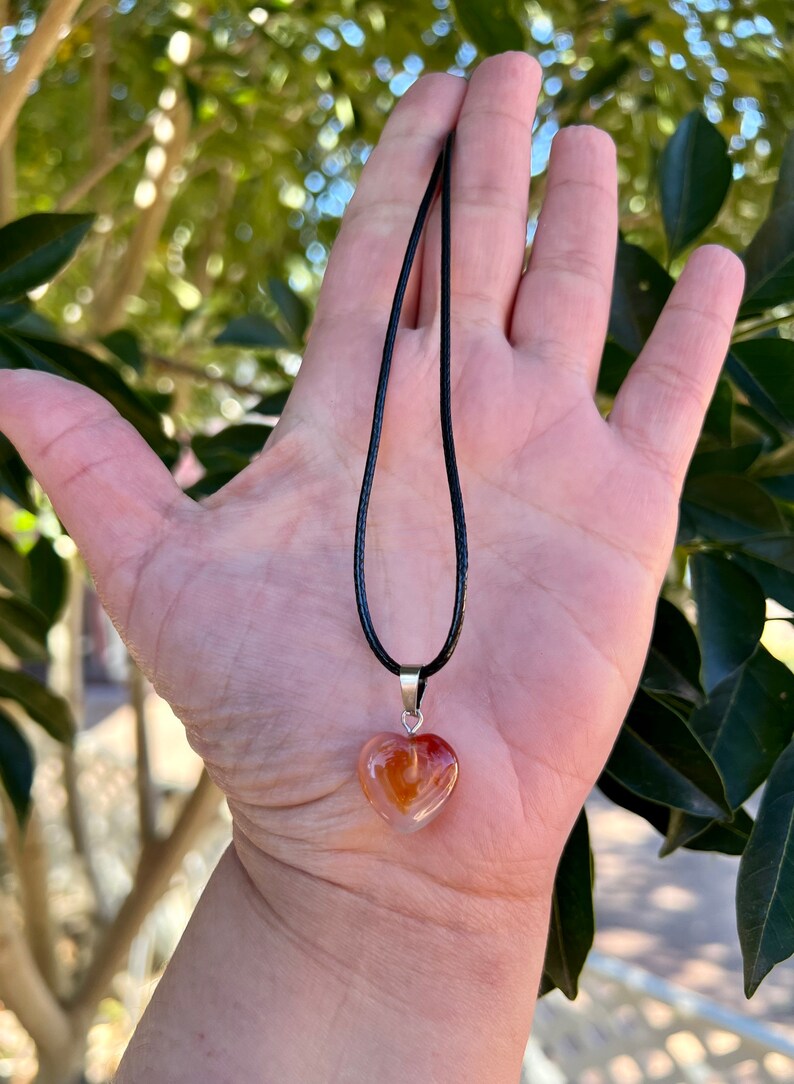 May include: A heart-shaped carnelian pendant necklace on a black cord. The pendant is a light orange color with darker orange and white streaks.