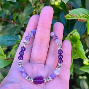May include: A purple amethyst bracelet with silver beads and a larger amethyst charm. The bracelet is made with different shades of purple amethyst beads.