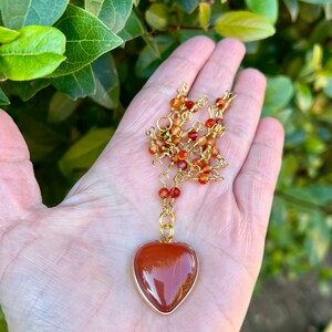 May include: A gold chain necklace with a heart-shaped carnelian pendant. The carnelian is a deep orange-red color and has a natural, striated pattern.