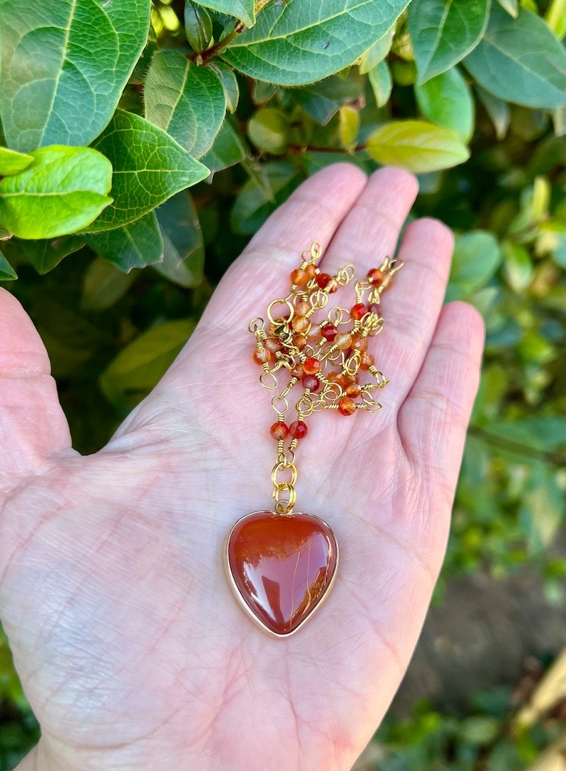May include: A gold chain necklace with a heart-shaped carnelian pendant. The carnelian is a deep orange-red color and has a natural, striated pattern.
