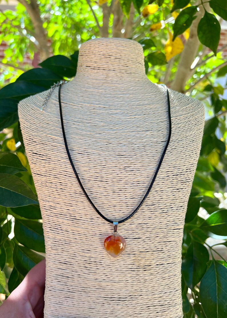 May include: A heart-shaped carnelian pendant necklace on a black cord. The pendant is a natural stone with orange, red, and white hues.
