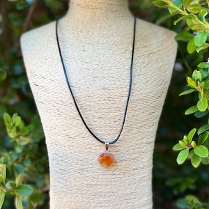 May include: A heart-shaped carnelian gemstone pendant on a black cord necklace. The pendant is a light orange color with darker orange and red streaks.