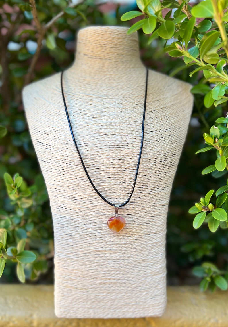 May include: A heart-shaped carnelian gemstone pendant on a black cord necklace. The pendant is a light orange color with darker orange and red streaks.