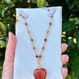 May include: A gold chain necklace with a heart-shaped carnelian pendant. The necklace is made with small carnelian beads and gold chain links.