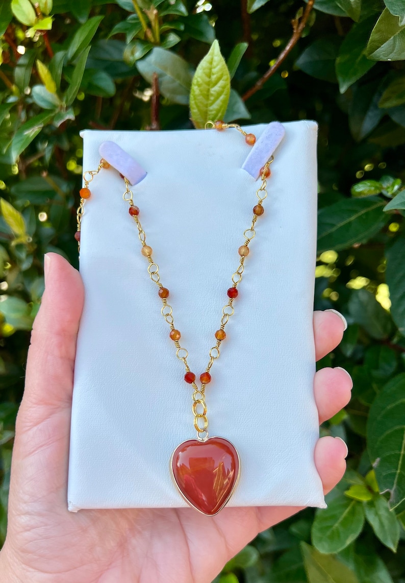 May include: A gold chain necklace with a heart-shaped carnelian pendant. The necklace is made with small carnelian beads and gold chain links.