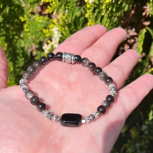 May include: A black and silver beaded bracelet with a black rectangular charm. The bracelet is made with small round beads and silver spacers.