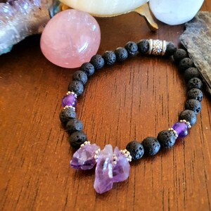 May include: A black lava rock bracelet with amethyst crystals and a silver charm. The bracelet is on a wooden surface.