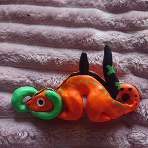 May include: Orange and green hand-painted clay figurine of a dragon with black accents. The dragon is curled up with its tail in its mouth.