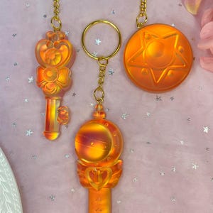 May include: Three orange and gold keychains featuring a key, a star, and a wand. The keychains are made of resin and have a shiny, iridescent finish.