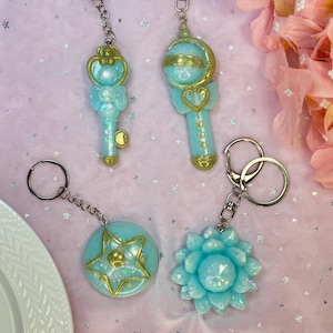 May include: Four keychains with silver rings. The keychains are light blue with gold accents. One is a key, one is a wand with a crescent moon, one is a star, and one is a flower. The background is pink with silver stars.