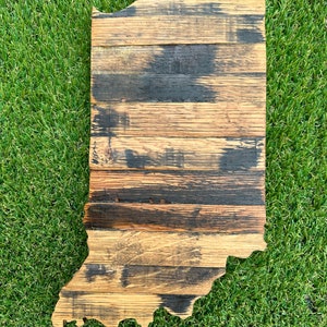 May include: A wooden state of Indiana outline, made from reclaimed bourbon barrel staves. The wood is a mix of light and dark brown with black char marks. The state is displayed on a green artificial grass background.